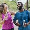 A woman and a man in athletic wear hold plastic water bottles as they walk in a park.