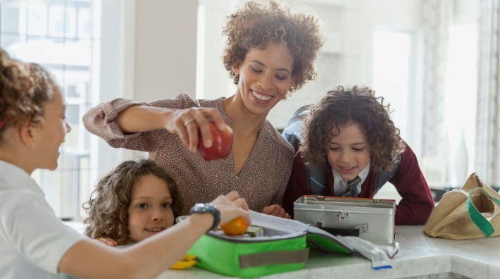 A woman holds an apple as a child puts an orange into a lunchbox on a counter and two kids watch. 