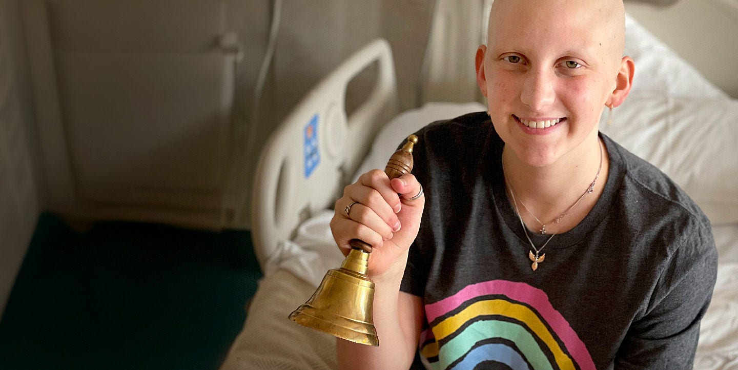 Persona sonriente sosteniendo una campana de bronce en una habitación de hospital.