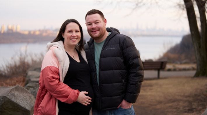 Cancer survivor and volunteer Zach Dane and his expecting wife smile in front of a lake on a cold day