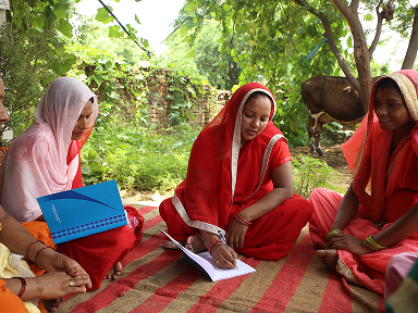 Several women sit outdoors on a mat under trees, reviewing papers and a booklet