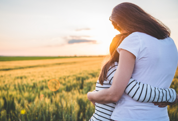 A mom and daughter hug while looking at a sunset across a field.