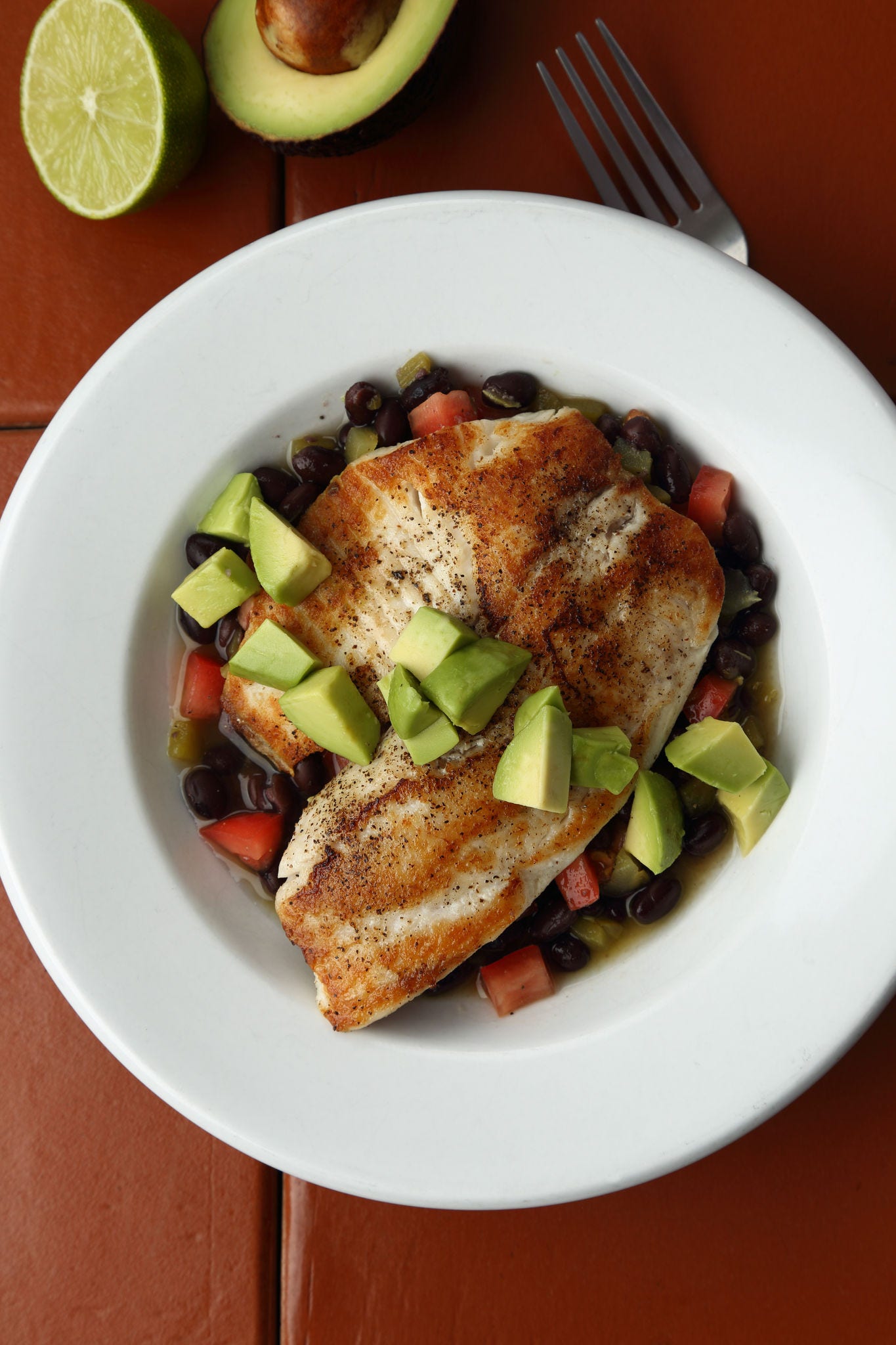 Chopped avocado sits on top of filets of tilapia over a mixture of black beans, tomatoes, and green chiles in a white bowl. 