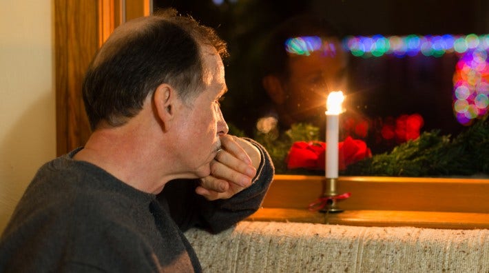 A depressed adult male looks out a window past Christmas decorations with Christmas lights in the background