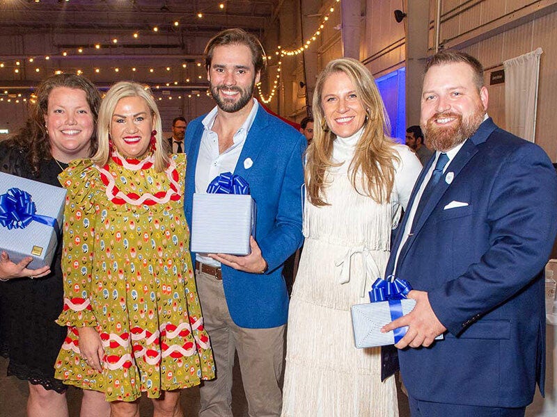 Five people holding gift boxes, smiling at indoor event.