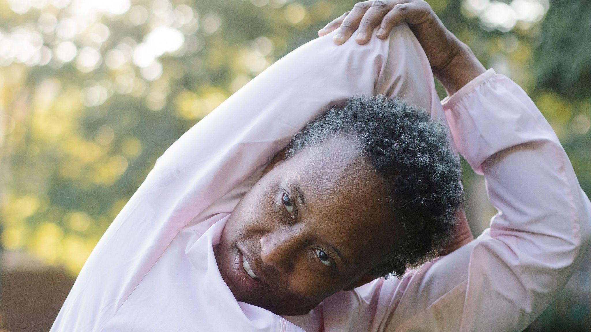 black woman with gray in her hair stretching wearing pink top
