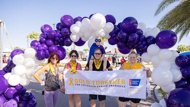 "Five people standing under a purple and white balloon arch, holding a 'Gold Together for Childhood Cancer Awareness' banner with the American Cancer Society logo