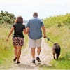 Seen from the back, a woman and a man walk a dog on a sandy seaside path with grasses and other plants on both sides. 