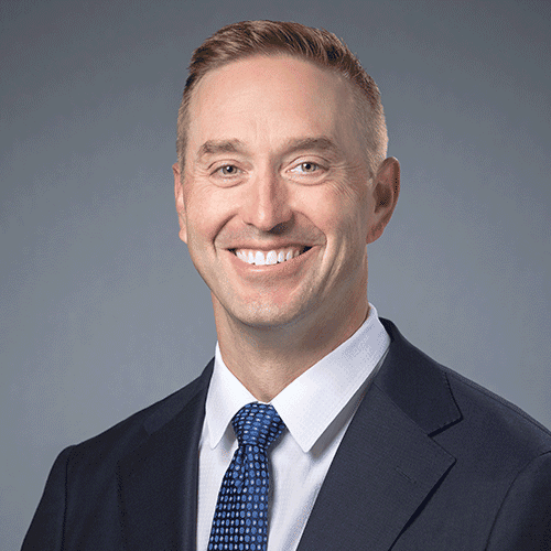 Smiling man in suit and blue tie, gray background.