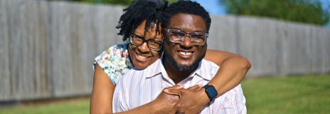 Una mujer que abraza a un hombre por detrás, sonriendo para una foto al aire libre