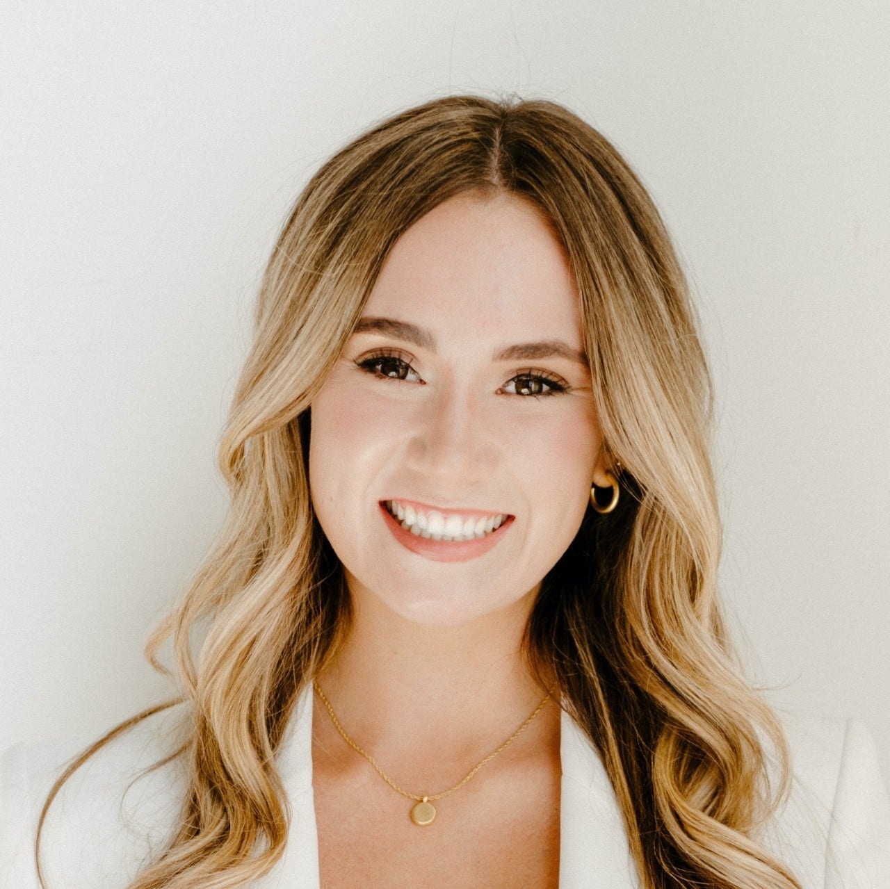 Young, white woman with long blonde hair parted in middle, in white jacket, with gold necklace and earring