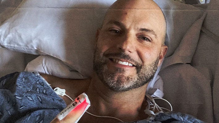 A Cancer patient (Jay) smiling in a hospital bed, wearing a gown and holding up a finger with a 'BIOPSY' bandage, surrounded by medical equipment.