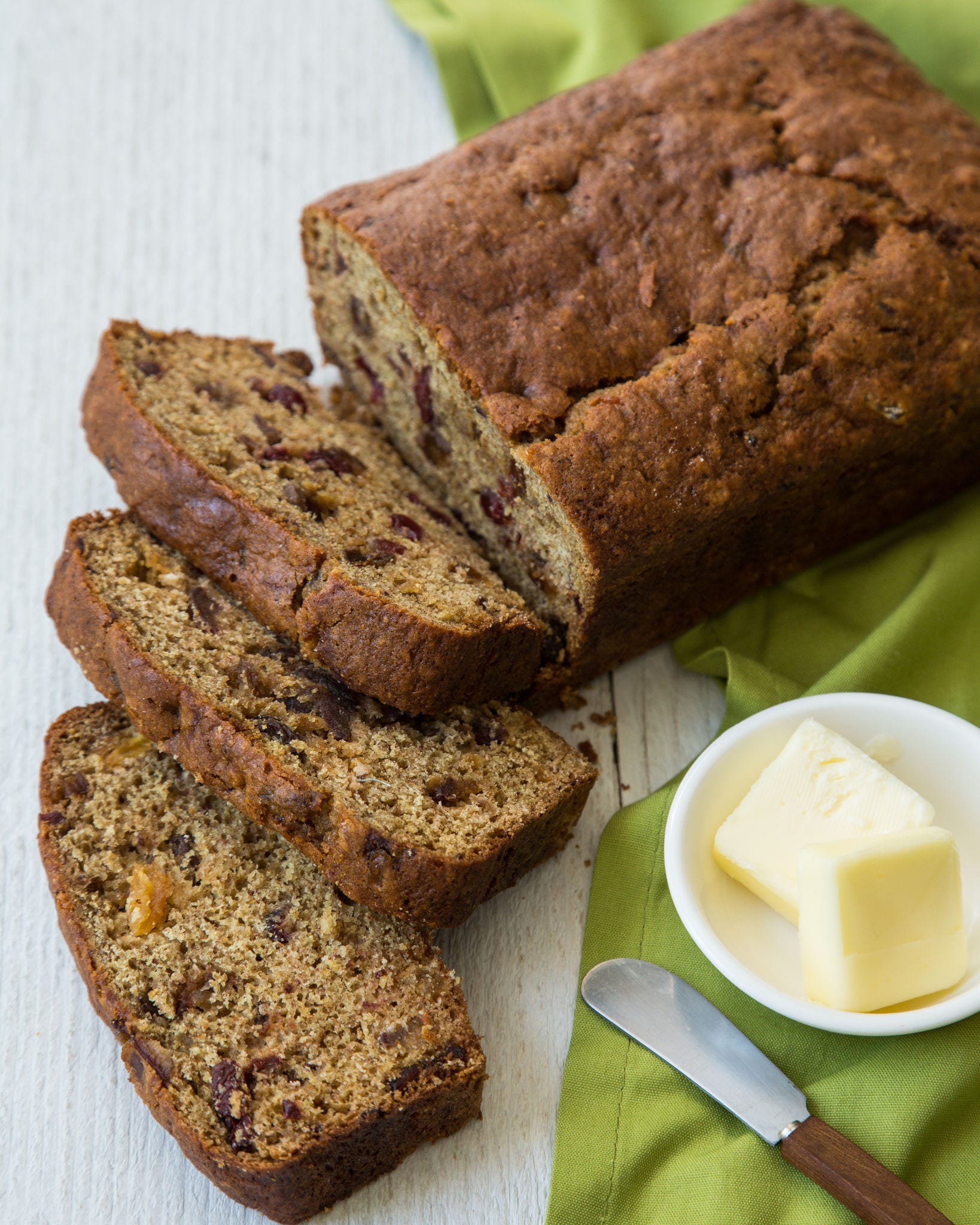Slices of a loaf of date and fruit bread lay next to a dish of butter and a spreading knife on a green cloth.