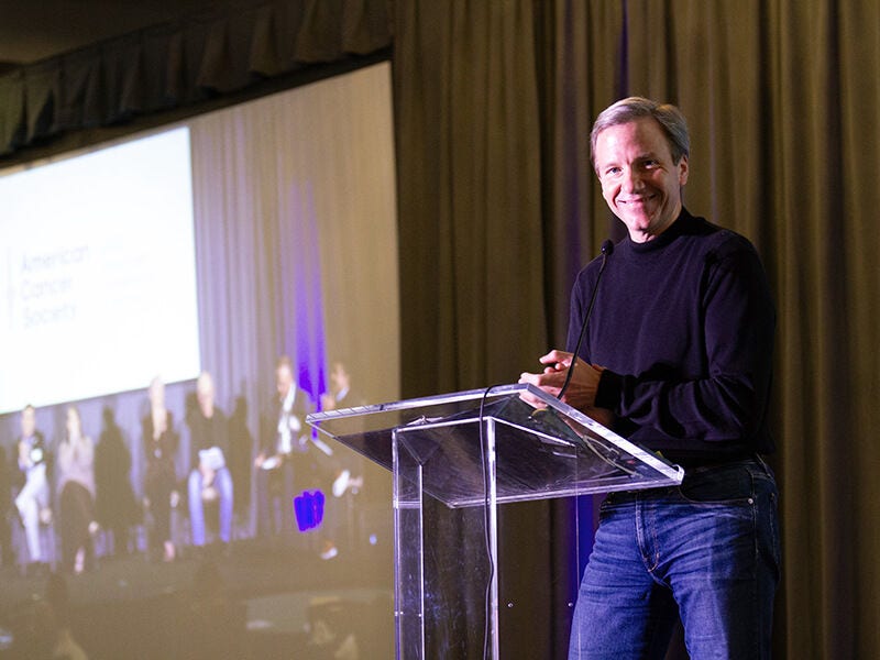 Man speaking at podium on stage, presentation in background.