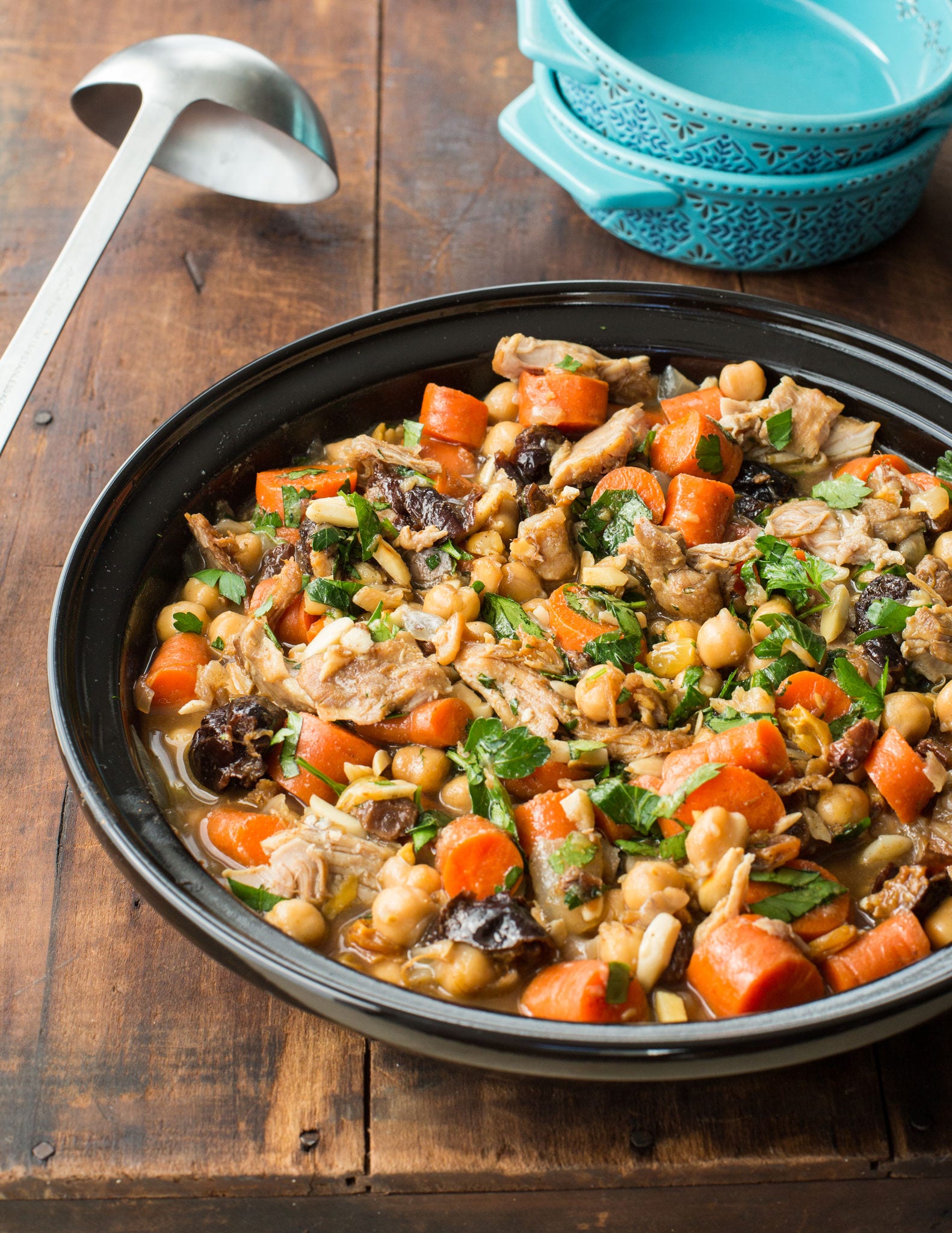 A ladle lays next to a large bowl of chicken tagine made with carrots, prunes, and chickpeas.
