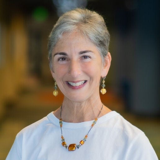 white woman, short gray hair, yellow bobble earrings white shirt with necklace