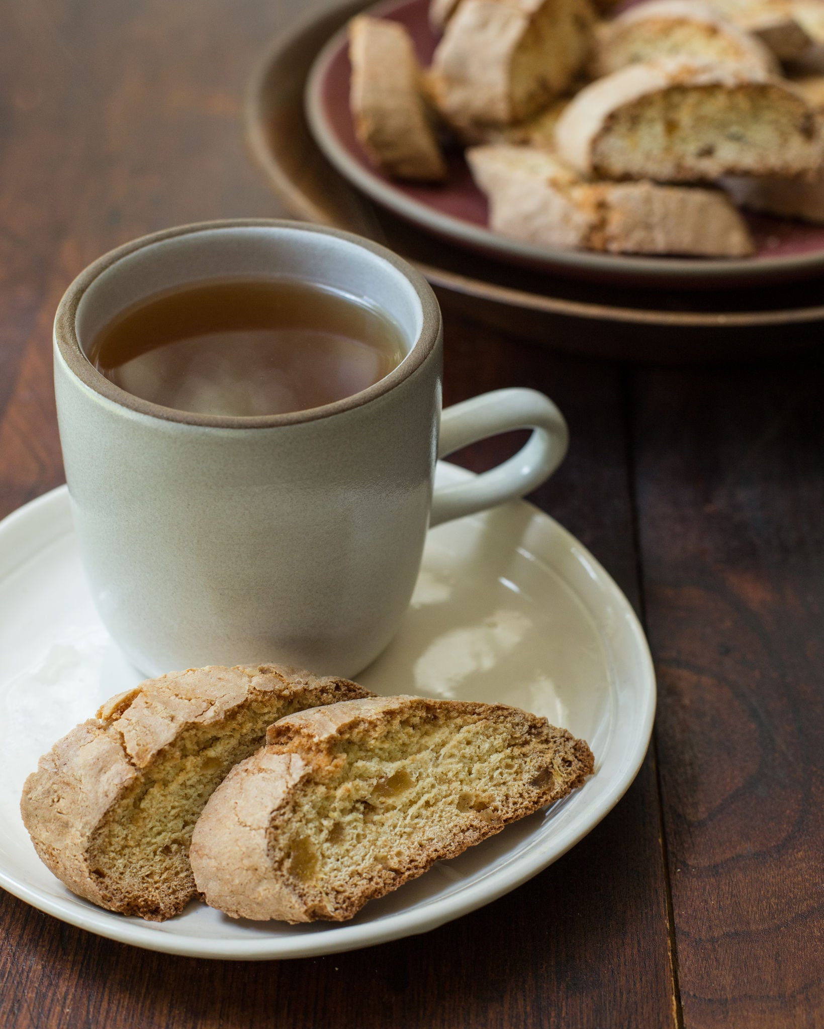 Two sliced pieces of biscotti lay on a white saucer next to a full mug of tea in front of a dish holding more biscotti slices.