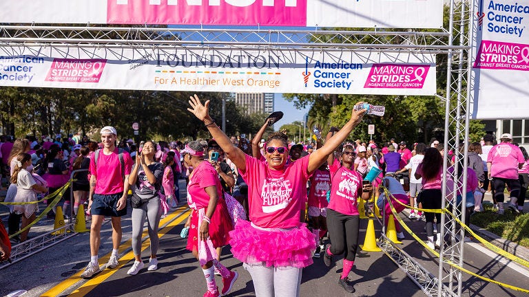 Participant in a pink shirt and tutu crosses the American Cancer Society Making Strides Against Breast Cancer finish line with arms raised.
