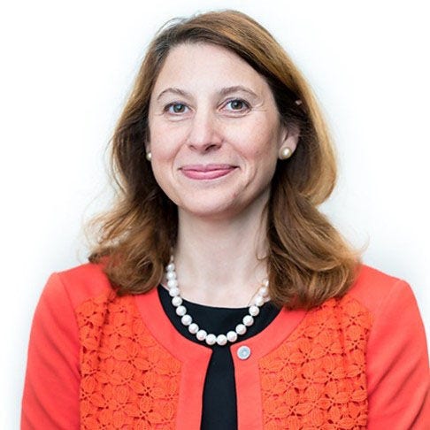 White woman smiling, long brown hair, pearl stud earrings, pearl necklace, orange sweater over a black blouse