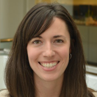 headshot of woman with brown hair, parted on right side, big smile showing teeth bit of office windows behind her