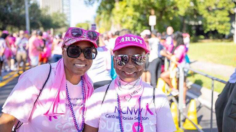 Two event participants wearing pink shirts, hats, and sunglasses pose together at a Making Strides Against Breast Cancer walk.