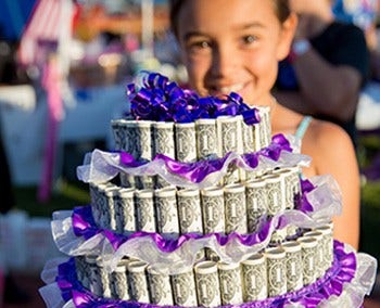 A young girl holding a birthday cake made out of rolled up dollar bills with purple ribbons on top.