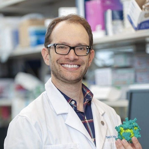 white man with red hair and beard, black glasses in blue plaid shirt with lab coat holding turquoise and bright green molecule