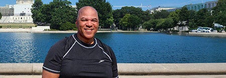 A person standing and smiling in front of a large reflecting pool with the U.S. Capitol building visible in the background on a sunny day.