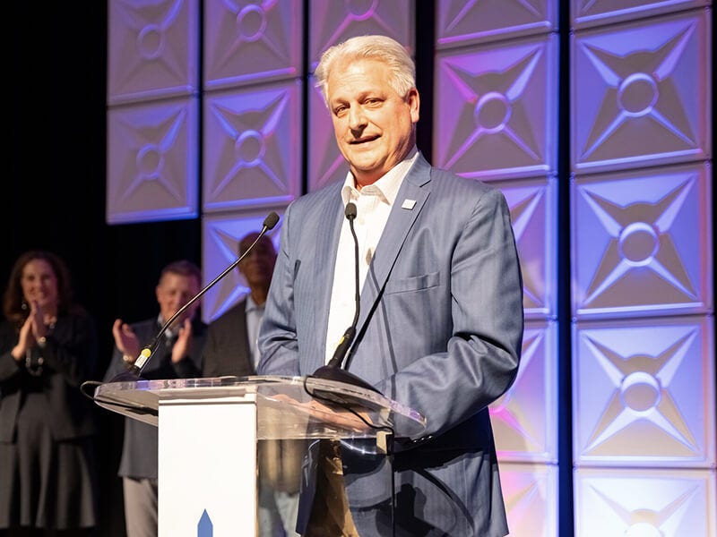 Man speaking at podium on stage, audience applauding behind.