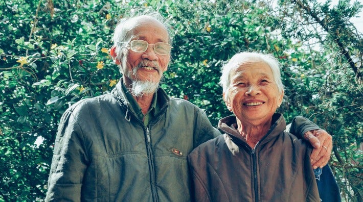 happy elderly Asian couple standing outside in front of greenery