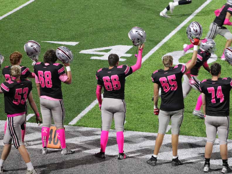 Young adult football players waving helmets and wearing jerseys with pink numbers and pink socks