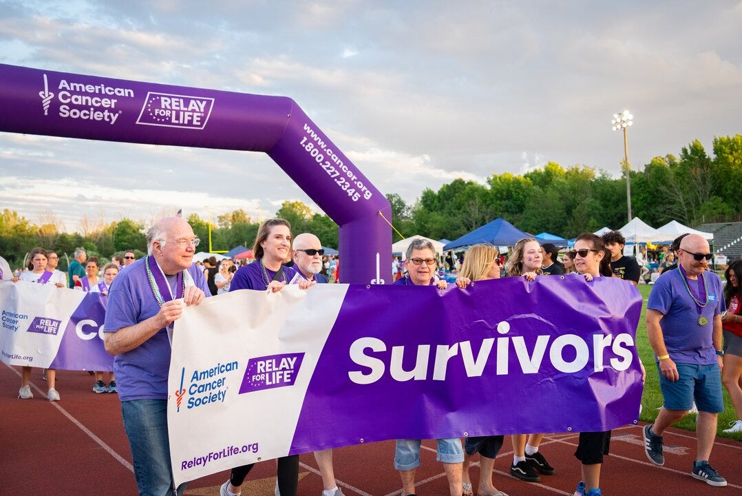 Group of people walking on a track holding a large purple “Survivors” banner at an American Cancer Society Relay for Life event.
