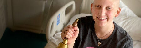 Persona sonriente sosteniendo una campana de bronce en una habitación de hospital.