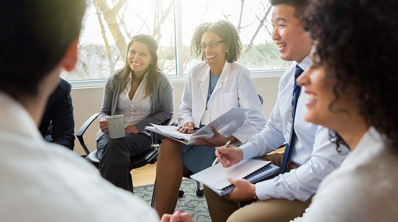 Diverse group of people sit in a circle in an office socializing with two women as the focus of the image