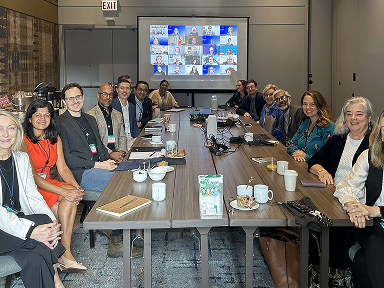 People sit around a long conference table during a hybrid meeting with participants visible on a screen.