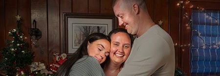 Three people hugging and smiling near a decorated Christmas tree.