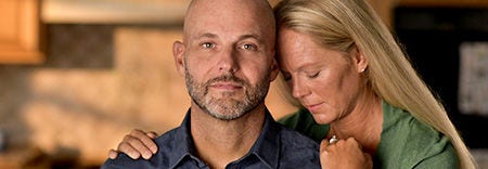 Man with a warm expression sitting close with a woman resting her head on his shoulder.