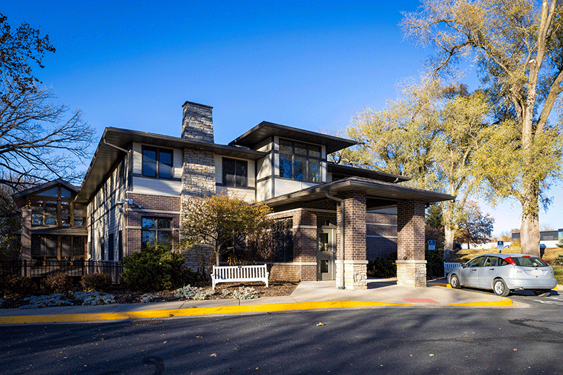 Brick building with covered entrance, bench, trees, and parked car.
