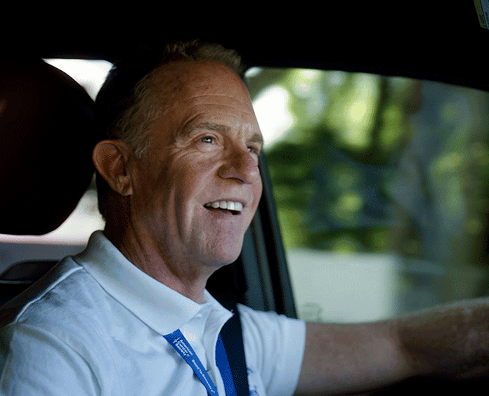 Smiling man driving car, wearing white shirt and lanyard.