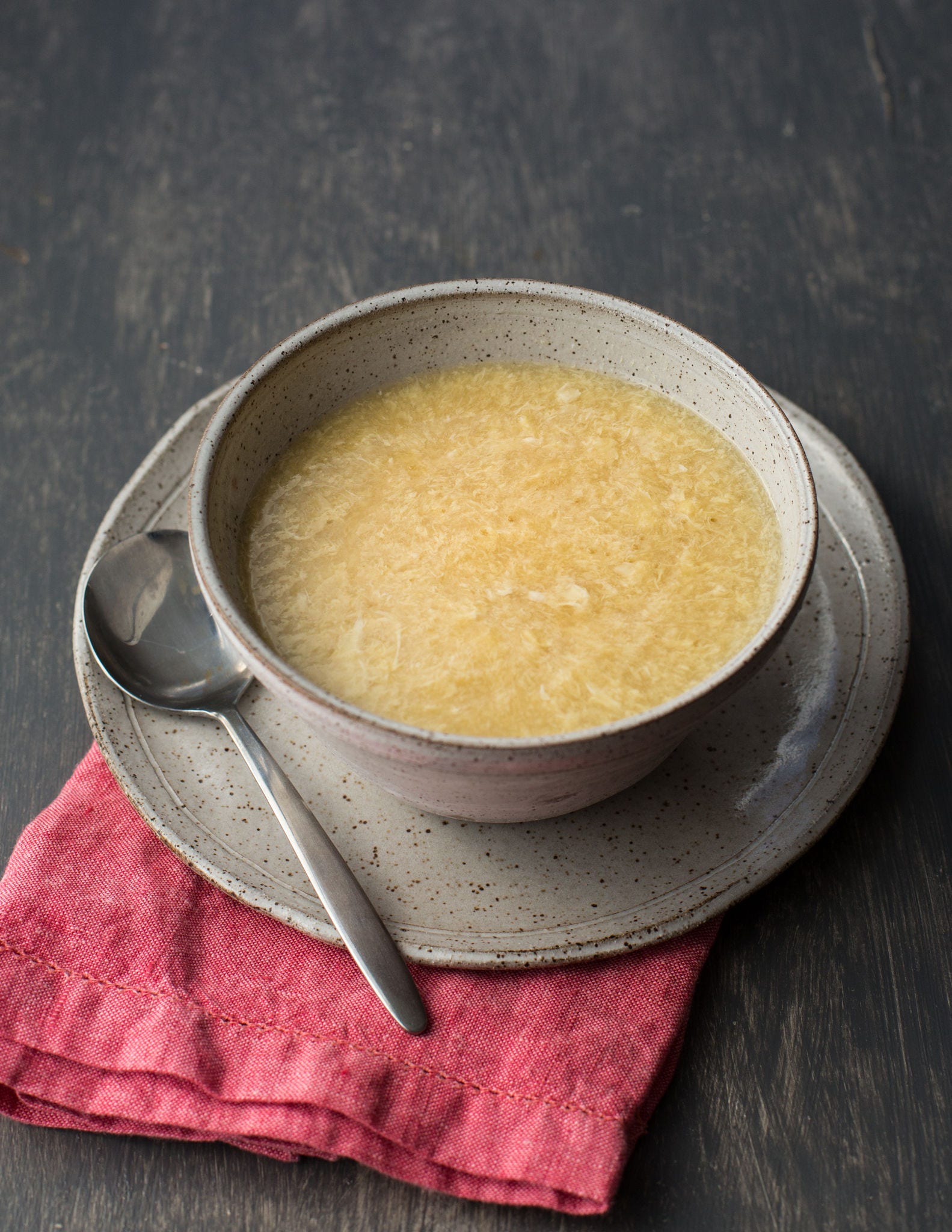 A stoneware bowl of egg drop soup rests on a stoneware plate on top of a folded pink cloth napkin.