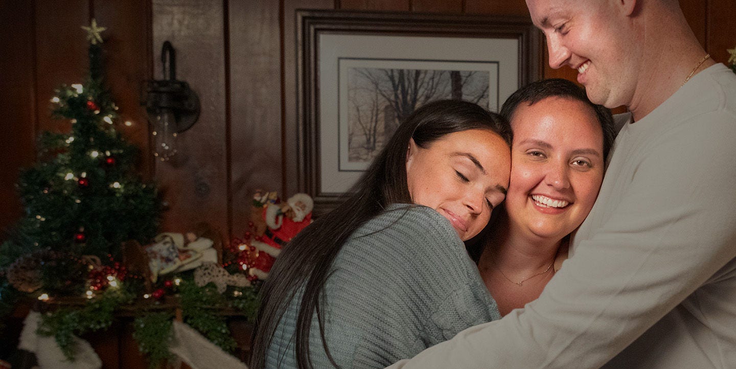 Tres personas abrazándose y sonriendo cerca de un árbol de Navidad decorado.