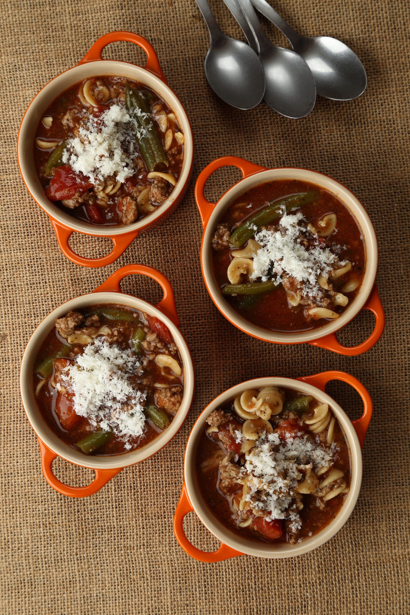 Four small bowls with orange handles, filled with a chunky turkey soup topped with grated Parmesan, sit on a piece of burlap.