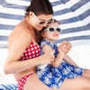 A woman in a red bikini with white polka dots puts sunscreen on a girl in her lap as they sit under a beach umbrella.