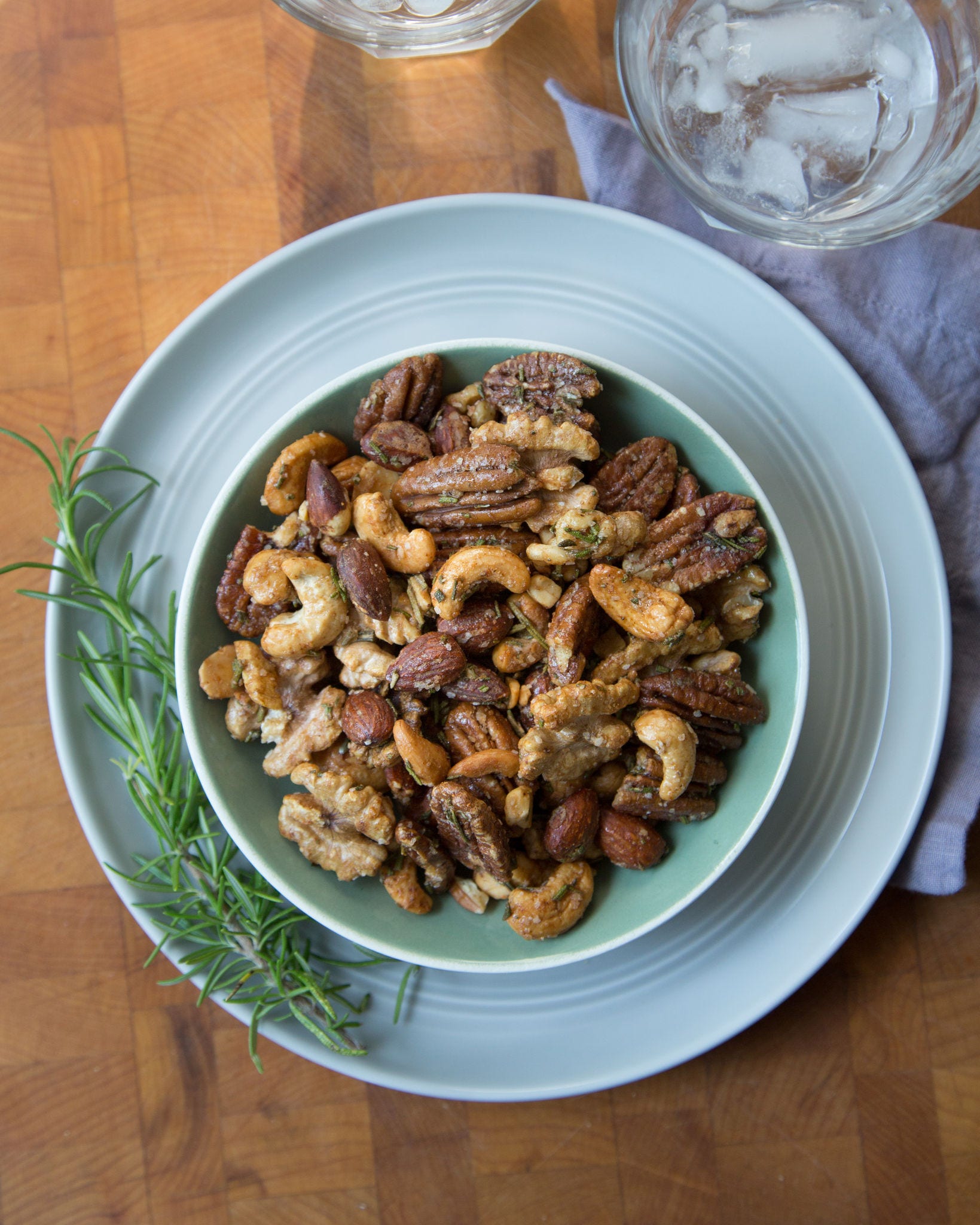 Seen from above, a bowl of sweet and spicy mixed nuts sits on a pastel blue plate next to a sprig of rosemary.
