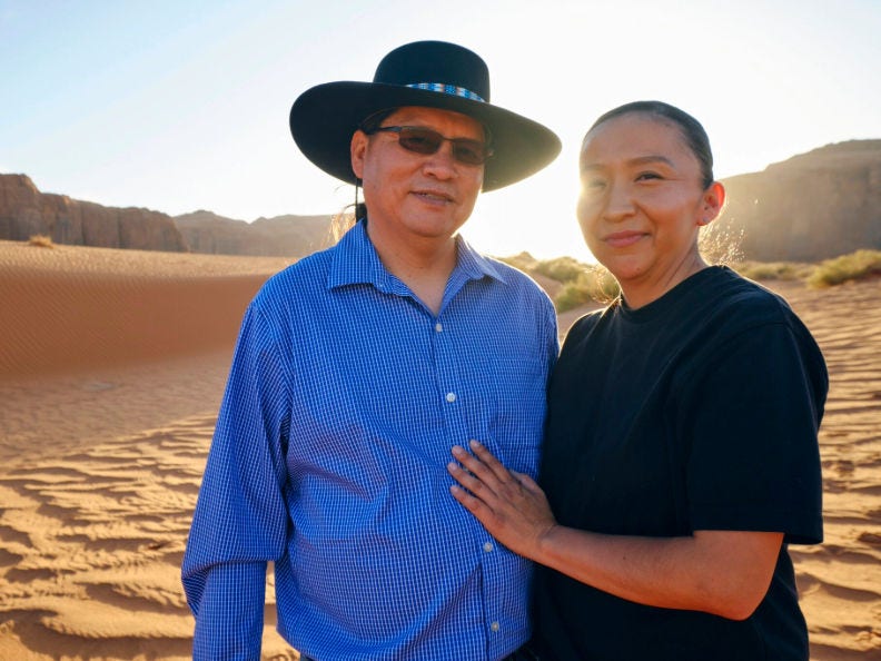 A Native American couple stand against a desert background with the sun behind them.