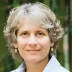 white woman with grayish hair wearing light blue collared shirt in front of blurred trees 