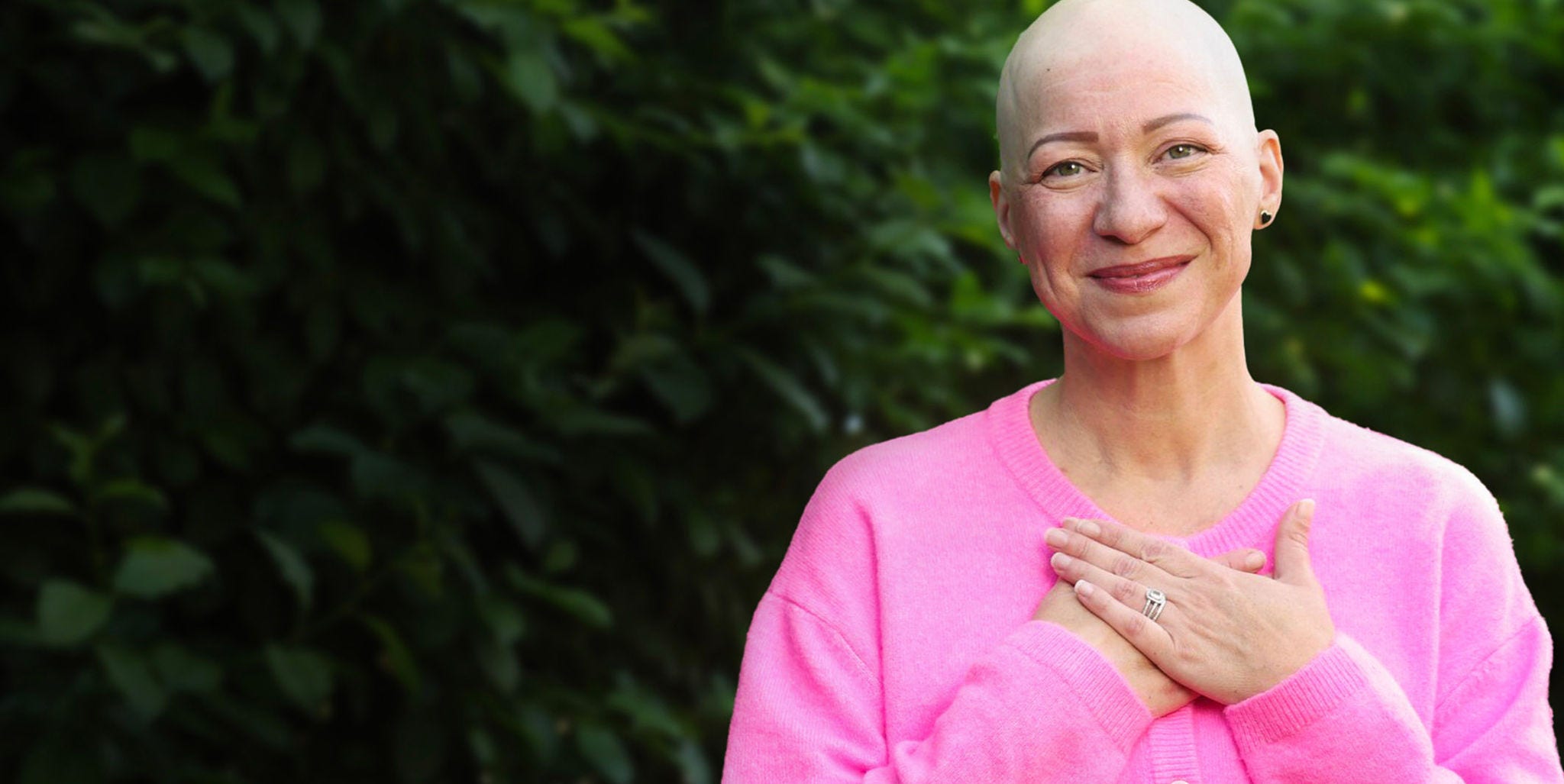 Person wearing a pink sweater, standing outdoors with hands over chest against a blurred green leafy background.