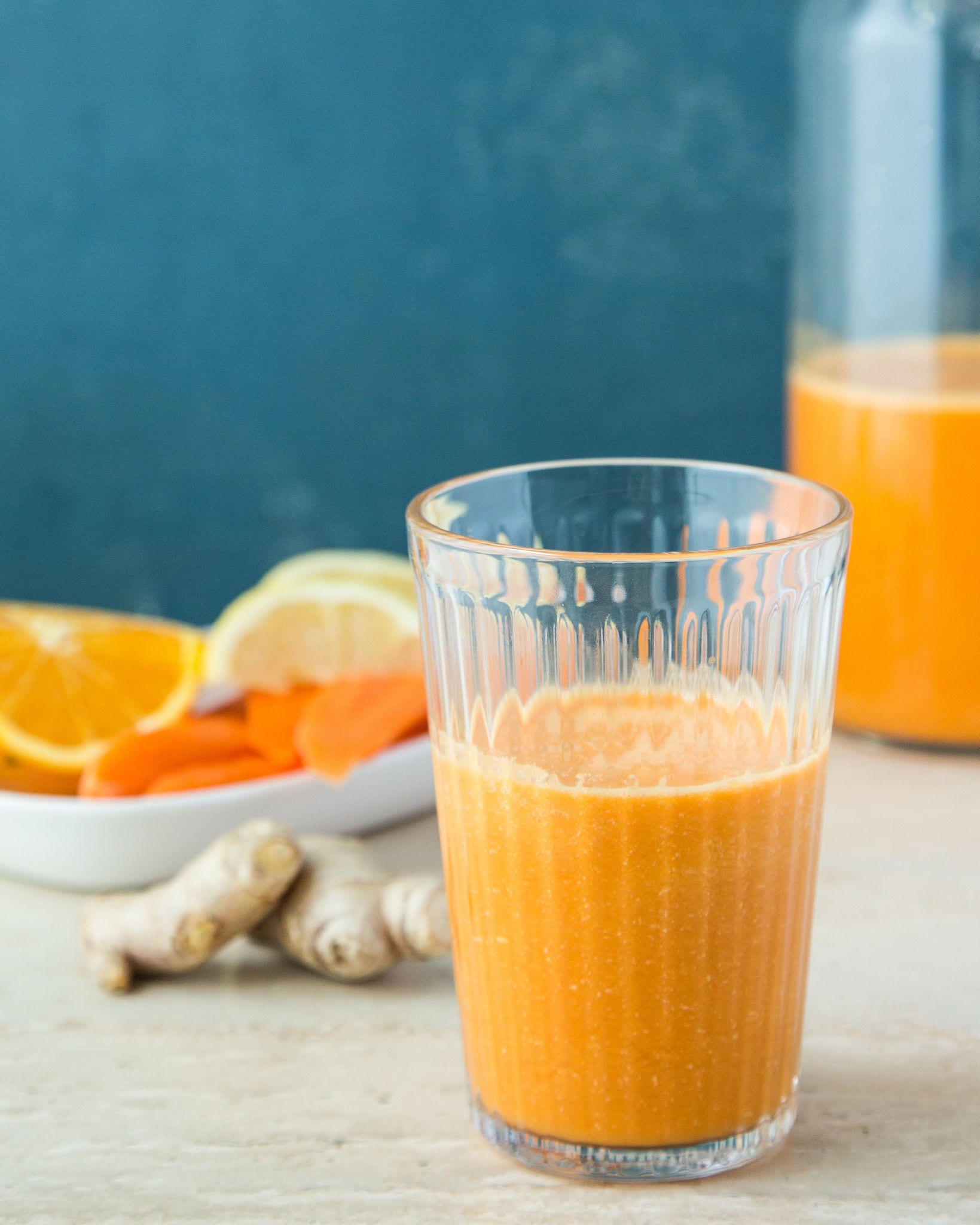 A glass of orange juice sits in front of a ginger root and a plate of sliced oranges and carrots against a blue background.
