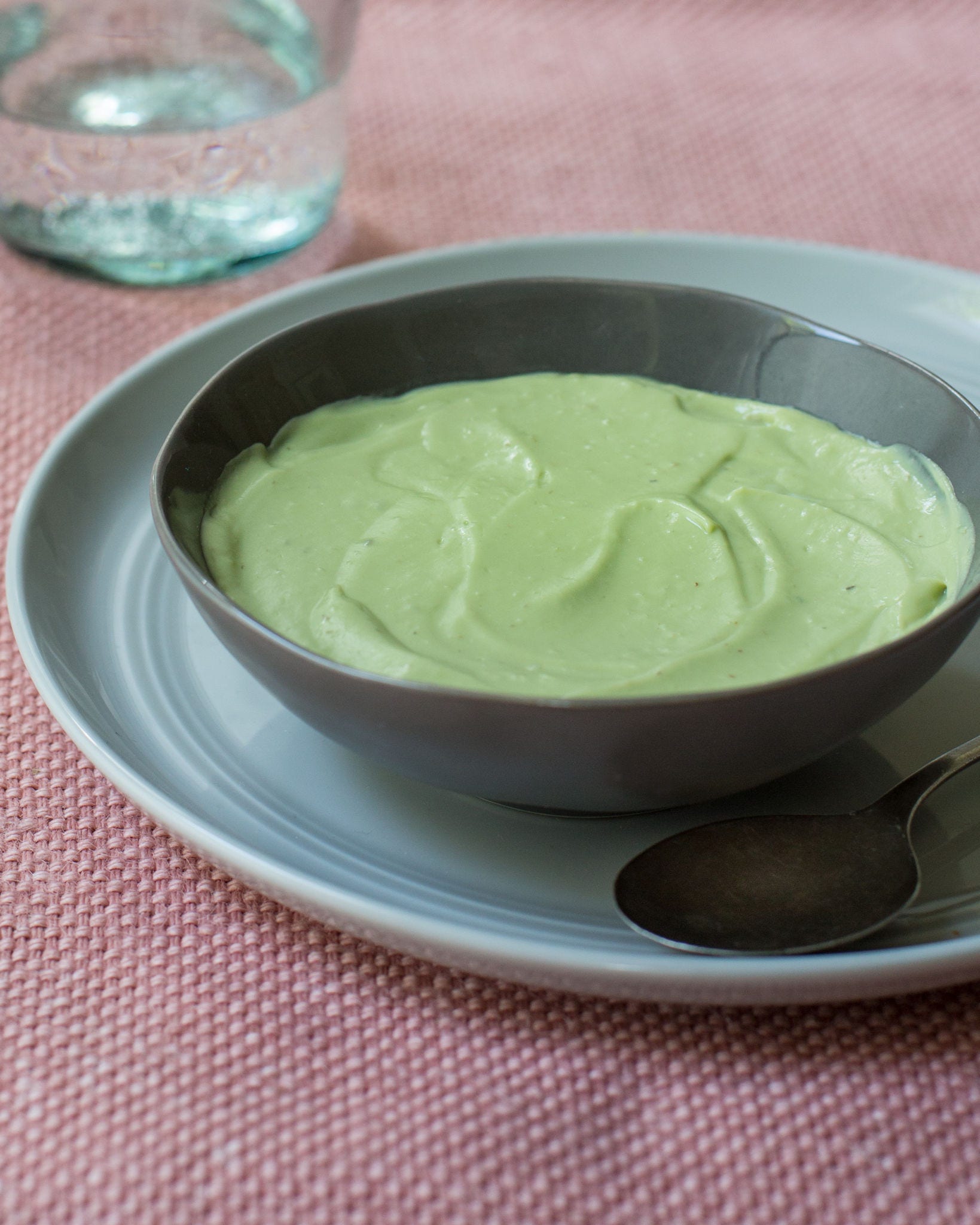 A blue-gray bowl of creamy pastel green soup rests on a blue-gray plate on top of a tablecloth.