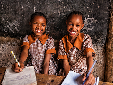 Two children in matching checkered school uniforms sit at a worn wooden desk, writing in notebooks against an old chalkboard wall.